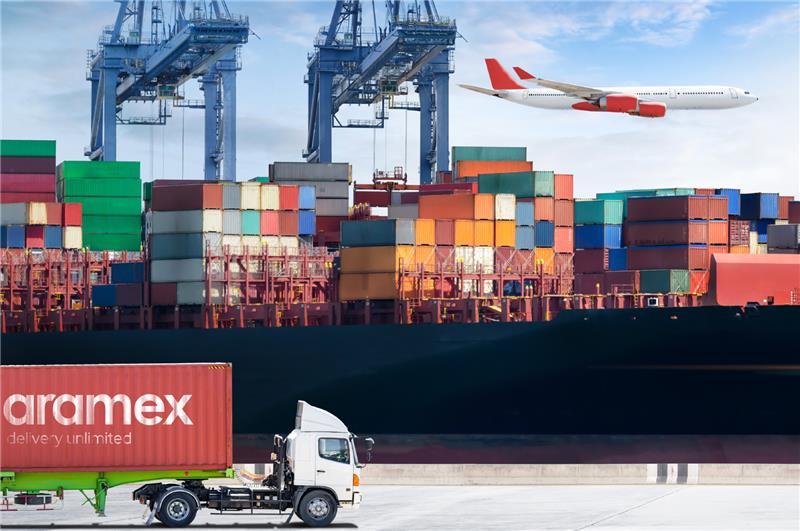 A shipping port with stacked colorful freight containers, a cargo ship, and a truck carrying an Aramex-branded container. A plane is flying in the background, representing global logistics and freight services.